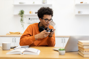 A young man with glasses is engrossed in a video game, holding a controller while sitting at a table with a laptop.