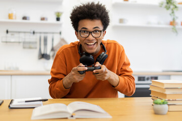 A young man with glasses is smiling while playing a video game, with headphones around his neck.