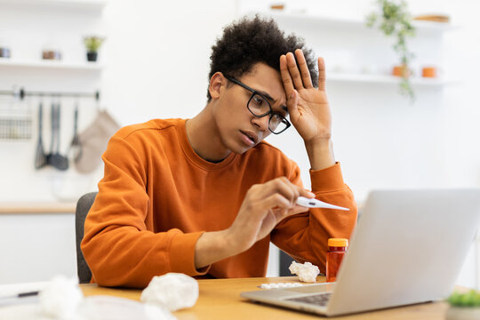 A young Black man with glasses checks his temperature while working on his laptop at home, appearing unwell.