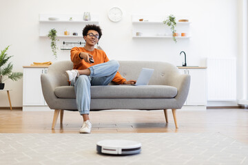 A young man relaxes on a sofa with a laptop and remote control while a robot vacuum cleans the floor.