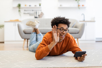 A young man with glasses lies on the floor, waving at his phone during a video call in a cozy living room.