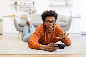 A young man with glasses lies on the floor, smiling and waving while holding a smartphone, likely engaged in a video call.