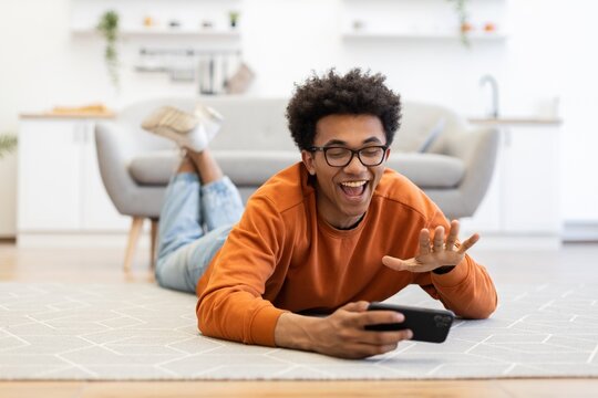 A young man with glasses lies on the floor, smiling and waving while holding a smartphone, likely engaged in a video call. - Powered by Adobe