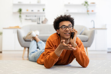 A young Black man wearing glasses lies on the floor, talking on his phone with a smile, in a cozy living room setting.