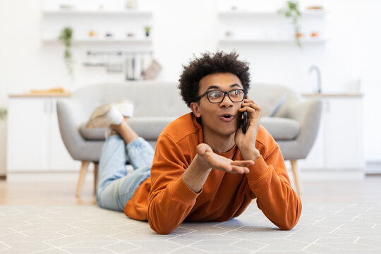 A young man with glasses lies on the floor, talking on his phone with an expressive gesture in a modern living room.