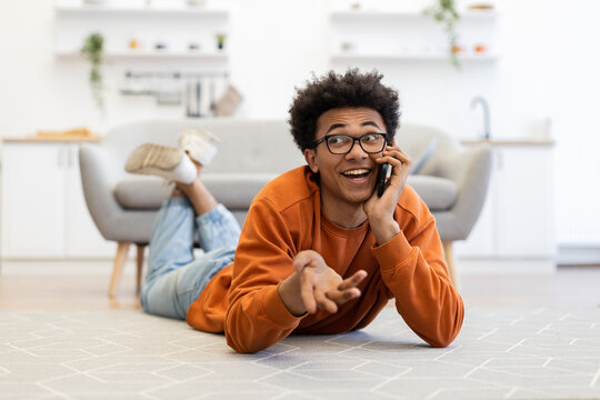A young man with glasses is lying on the floor, talking on his phone with a surprised expression, in a cozy living room.