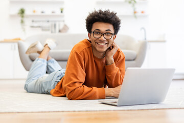 A young man with glasses smiles while using a laptop, lying on the floor in a cozy living room setting.