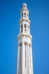 A tall, elegant white mosque minaret rising against a bright blue sky. The tower features arched windows, subtle geometric detailing, and traditional Islamic architectural elements