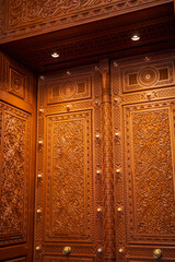 A detailed close-up of an ornate wooden door featuring intricate Islamic carvings, geometric motifs, floral patterns, and decorative brass accents. The craftsmanship showcases traditional Middle East