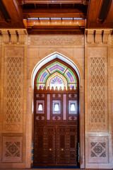 A beautifully detailed interior scene showcasing an ornate wooden door topped with a vibrant stained-glass arch in traditional Islamic style. The surrounding walls feature carved geometric patterns