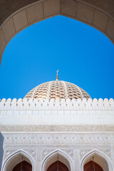 A beautifully composed exterior view of a white mosque dome topped with a crescent moon finial, framed elegantly by an Islamic arch. The architecture features intricate carvings, geometric patterns