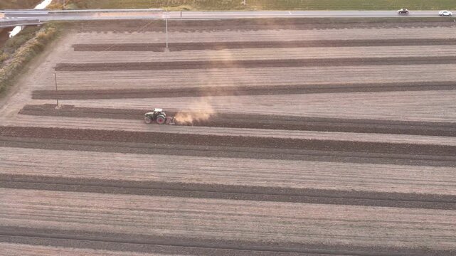 Tractor conducting deep tillage across structured rows in a dry Po Valley field, with visible dust plume and access road, near San Pietro al Cerro, PC, Italy