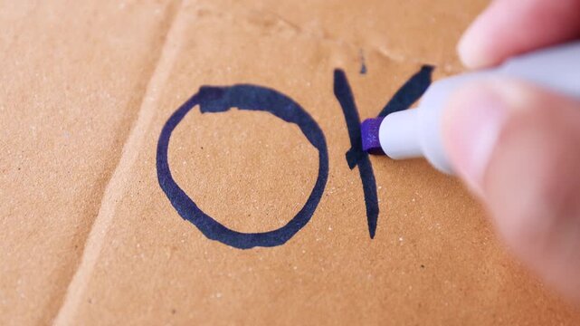 Hand writing "OK" with blue marker on cardboard, symbolizing approval, correctness, or a simple positive message, close-up