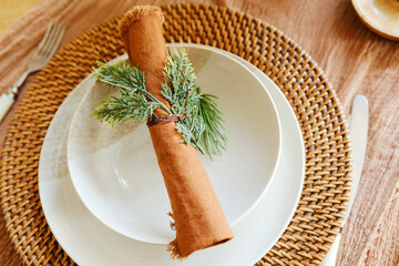 Warm table setting with woven placemat white plate brown napkin green pine sprigs detail