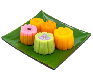 Colorful, flower-shaped desserts rest on a green banana leaf, arranged on a square, dark green platter. The background is a stark black