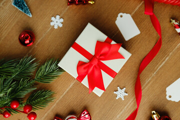 Gift box with red ribbon surrounded by pine needles, ornaments, snowflakes and empty tags on wood