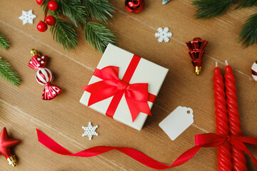Festive Christmas gift box with red ribbon, pine sprigs, ornaments, and candles on wooden background.