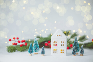 Miniature winter village scene with ceramic house, evergreen trees, red berries, and soft lights.