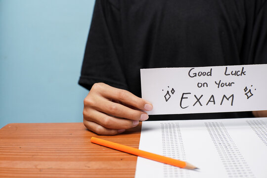 A student holding a handwritten good luck exam card above a multiple-choice test sheet and pencil, symbolizing study, motivation, and exam preparation.
