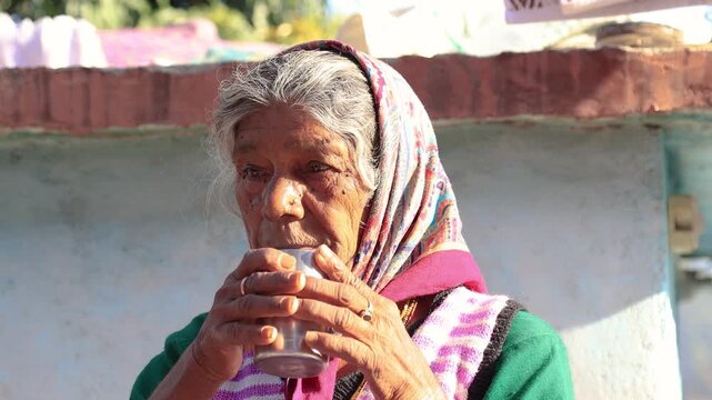 Old Indian rural woman around 80 to 85 years enjoying a warm cup of tea outdoors at her village home, sitting peacefully in a natural mountain setting.