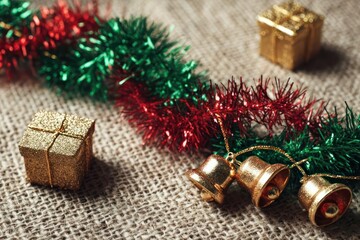 Festive christmas decorations including shiny tinsel garland golden gift boxes and jingle bells on a textured burlap background