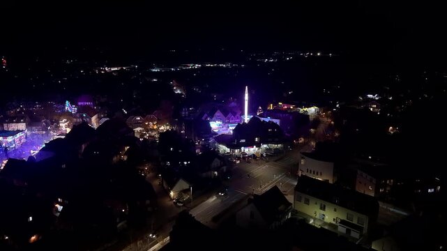 Carnival funfair event in american town with lighting tower and fairground rides. Driving cars on main street at night. Aerial flyover shot.