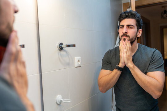 Young man touching beard, looking at his reflection in bathroom mirror, thinking about grooming and self-care routine