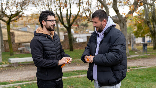 Two men in casual outfits standing in a park, peeling oranges and having a conversation during autumn