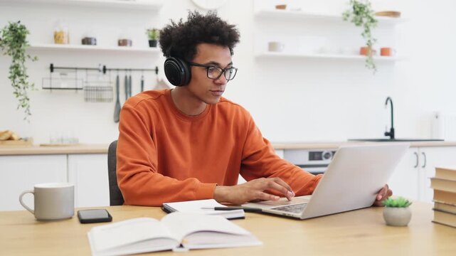 A young man with glasses listens to music on headphones while working from home in his kitchen.