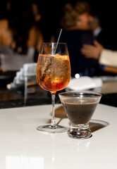 Stylish bar setting with two cocktails on a glossy counter an Aperol Spritz with ice and a dark espresso cocktail. Elegant party atmosphere with blurred guests in the background