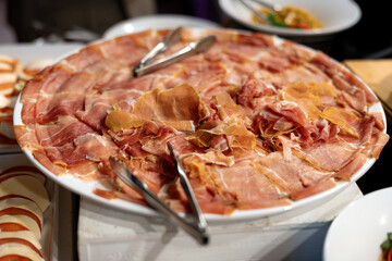 Large platter of thinly sliced prosciutto arranged for a buffet table. Fresh, savory cured meat displayed with serving tongs, creating an appetizing presentation for events or catering