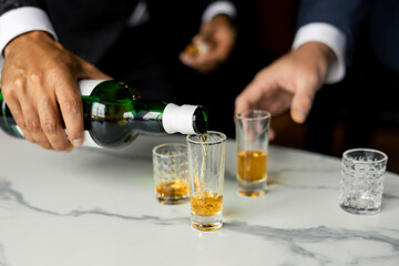 Close-up of a man pouring whiskey from a green bottle into shot glasses on a marble table. Elegant celebration moment, featuring hands in formal suits and golden liquor