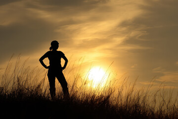 Silhouette of person standing confidently on grassy hill against dramatic sunset sky, evoking sense of empowerment and tranquility