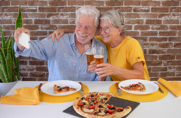 Happy senior couple looking at phone taking a selfie while toasting with a glass of beer.. Elderly woman and man share a traditional Italian pizza for dinner