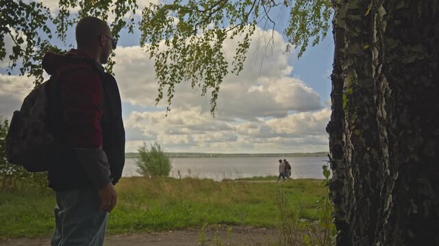 A man stands, contemplating the lake and the people walking along the shore.