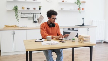 A young man with glasses smiles while looking at his laptop and holding a mug of coffee in a bright kitchen setting.