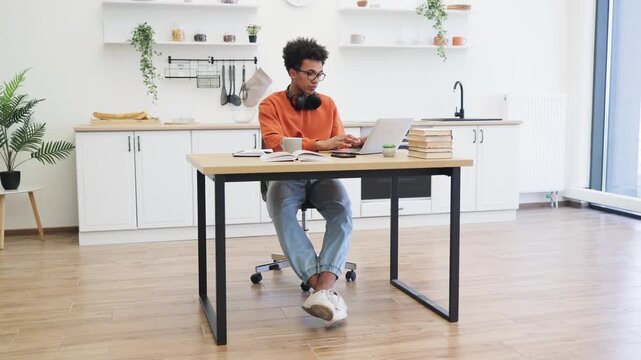A young man with glasses enjoys a cup of coffee while working on his laptop at a table in his kitchen.