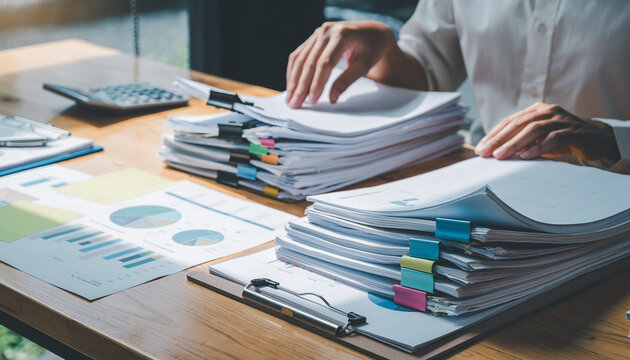Office desk with stacked paperwork, colorful binder clips, hand sorting documents, financial charts, and calculator, creating busy and organized work environment - Powered by Adobe