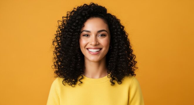 Happy woman with curly hair smiling in a yellow sweater against an orange background.