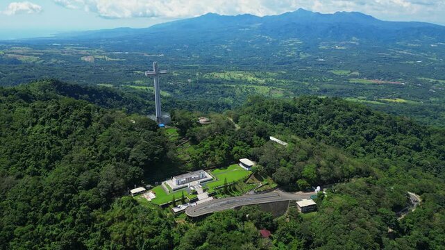 Slow push aerial highlighting the towering Mt. Samat National Shrine, gradually revealing its cross monument framed by lush forested hills and sweeping valleys in Pilar, Bataan, Philippines.