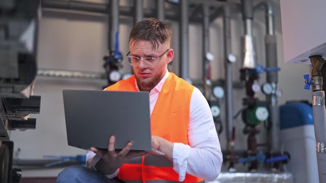 Professional plumber in safety vest diligently works on laptop, analyzing data in industrial setting. Appears concerned while troubleshooting complex system, focused on resolving technical issues.