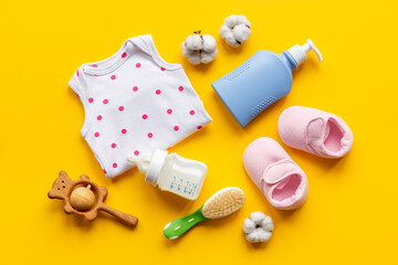 Baby bodysuits and eco friendly accessories - booties, wooden rattle toy and baby bottle with milk on yellow background, top view