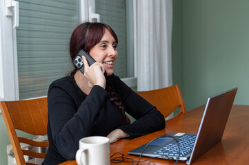 Smiling woman working remotely from home, talking on phone with laptop and coffee on wooden table
