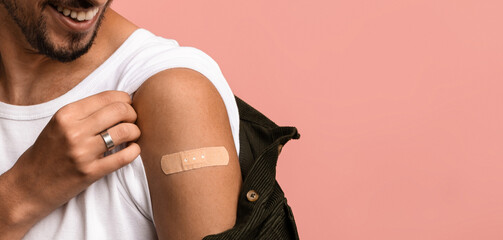 A man smiles while gently pulling up his sleeve to reveal a bandage on his arm. He has just received a vaccination in a light-colored studio, with a pink background adding warmth.