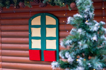 Wooden Christmas hut wall with decorative green-yellow window, red boxes, festive garland with pinecones, and a snowy artificial Christmas tree.