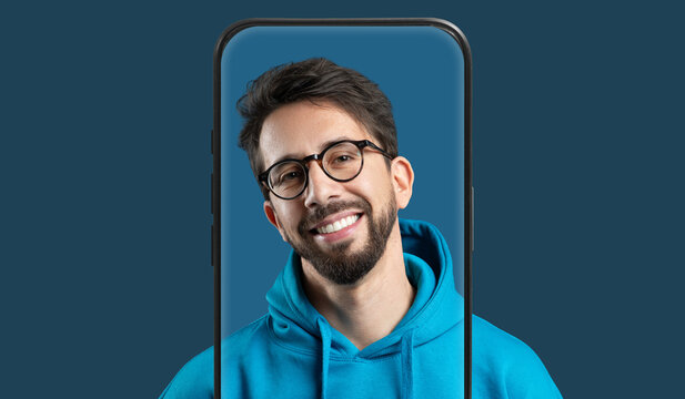 A man with glasses and a beard smiles brightly while wearing a blue hoodie. His image is creatively displayed within the frame of a smartphone, against a solid dark background. - Powered by Adobe