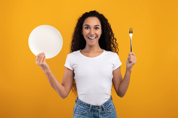 Smiling young lady with curly hair holds a fork in one hand and a plate in the other. She looks thrilled to eat, posing against a vibrant yellow background.