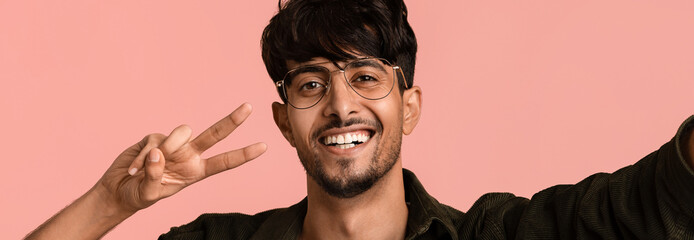 A young man wearing glasses smiles brightly while giving a peace sign with his hand against a soft pink background. He is dressed casually and exudes a cheerful and friendly vibe.
