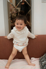 Fototapeta premium Happy Asian toddler girl playing and standing on a sofa at home, laughing and having fun.