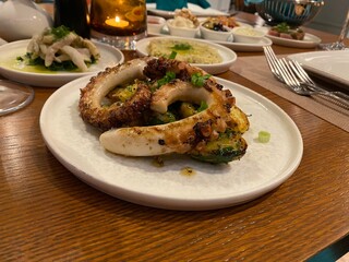 A delicious serving of grilled octopus tentacles and roasted potatoes on a white plate, with other appetizers and a lit candle on a wooden restaurant